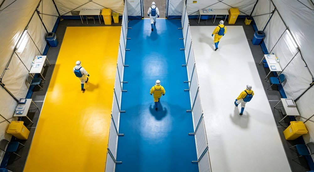 Aerial top down view of decontamination tent interior showing three zone flow layout with colored floor sections in yellow blue and white and workers walking through