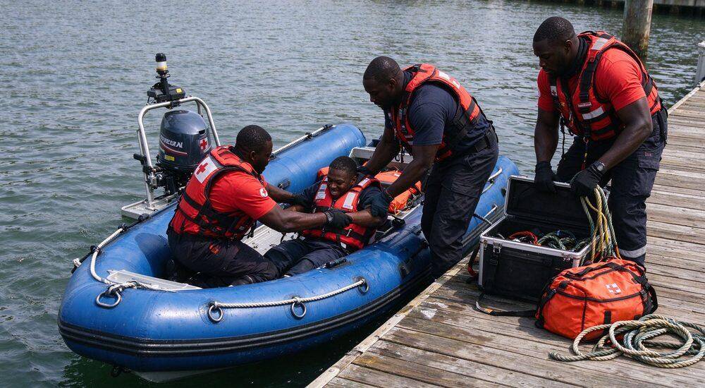 Boarding and loading operation where male rescuer stabilizes inflatable rescue boat on shore while injured victim climbs aboard showing patient transfer procedure for flood response boat Boarding and loading operation where male rescuer stabilizes inflatable rescue boat on shore while injured victim climbs aboard showing patient transfer procedure for flood response boat